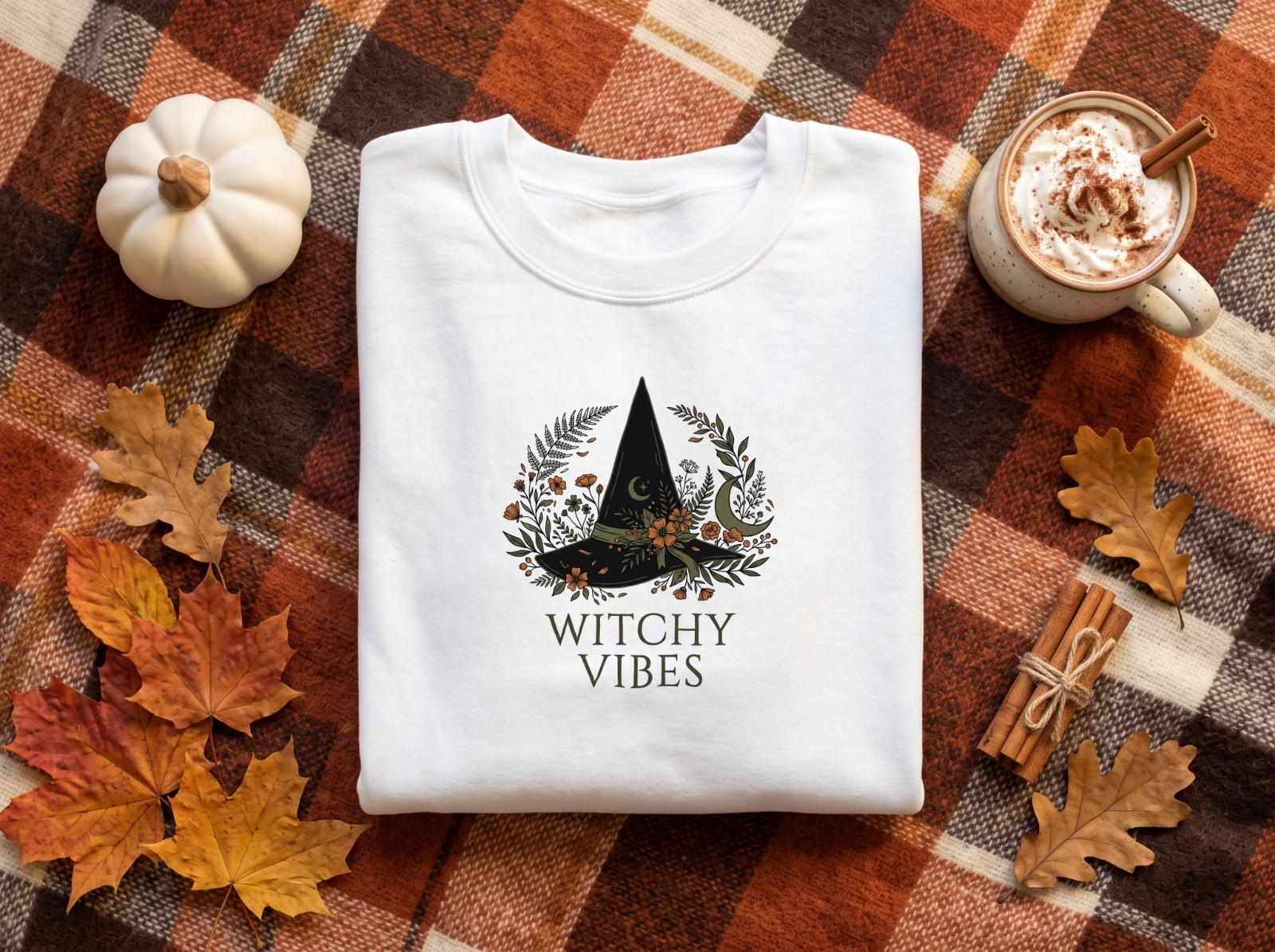 Folded White Sweatshirt Halloween on a Plaid Blanket showing a Witchy Vibes botanical witch hat design