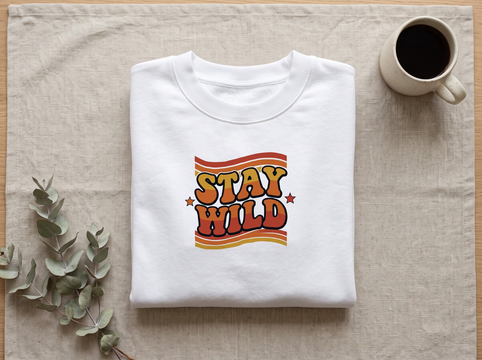 Folded White Sweatshirt on Linen with Eucalyptus showing the retro Stay Wild design