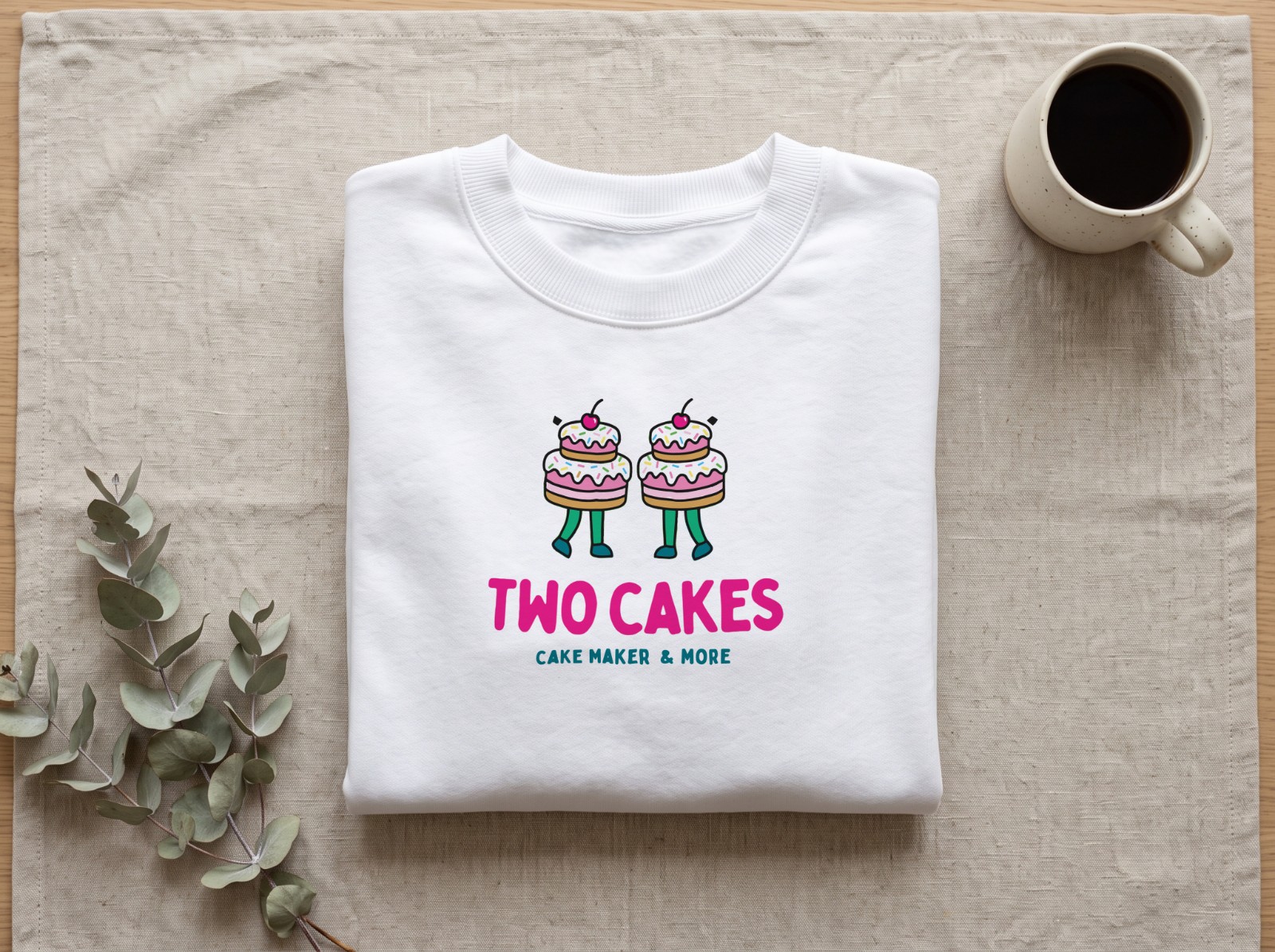 Folded White Sweatshirt on Linen with Eucalyptus showing the Two Cakes logo