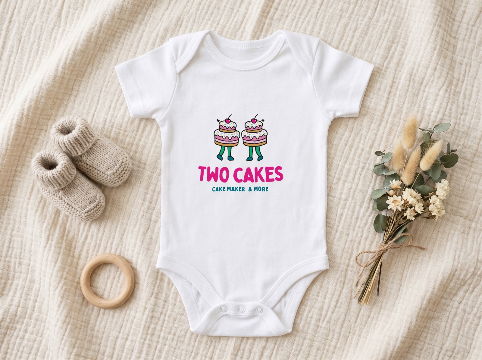 White Baby Onesie Flat Lay on a Muslin Blanket showing the Two Cakes logo