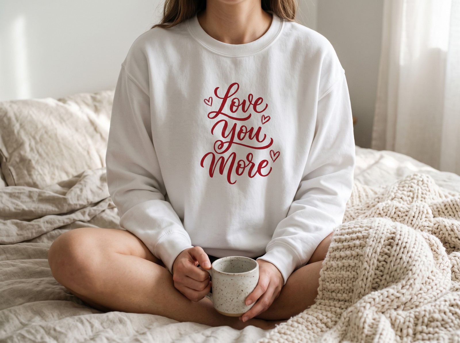 White Sweatshirt on a Woman Sitting on a Cosy Bed showing a Love You More lettering