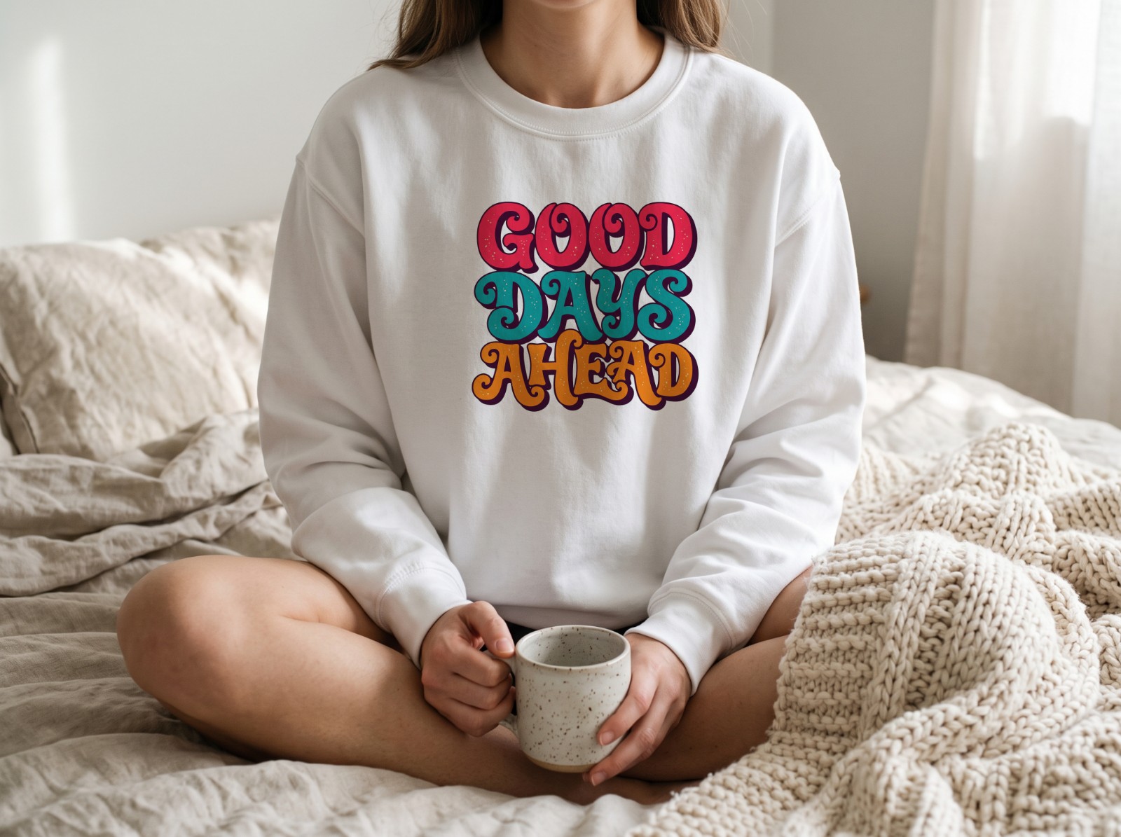 White Sweatshirt on a Woman Sitting on a Cosy Bed showing a retro Good Days Ahead lettering